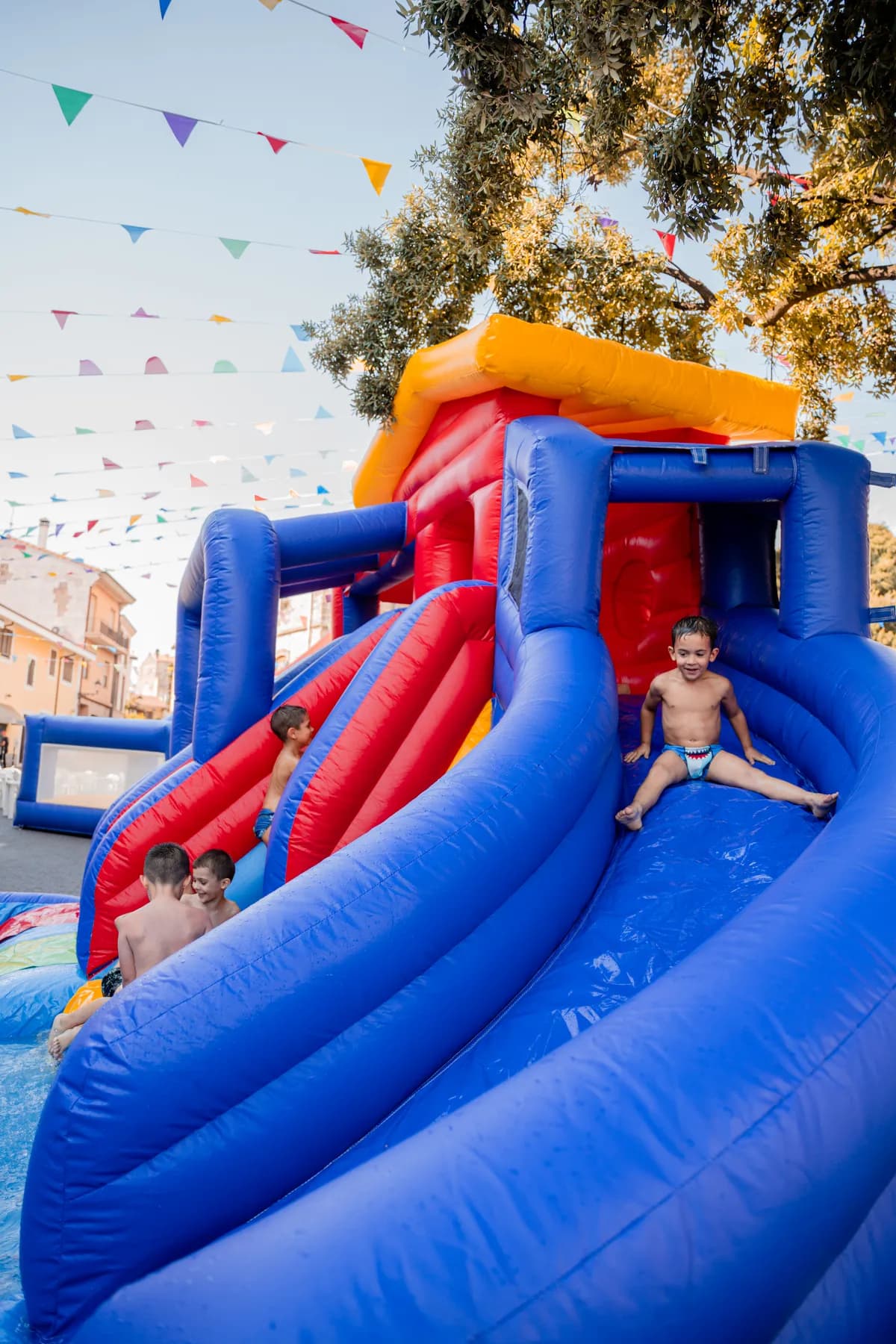 Bambini piccoli giocano sullo scivolo gonfiabile a casetta dell'area baby, sotto le bandierine della piazza