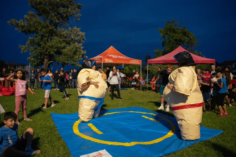 Due persone dentro i costumi gonfiabili da sumo che si sfidano sul tappeto, coi bambini intorno che guardano
